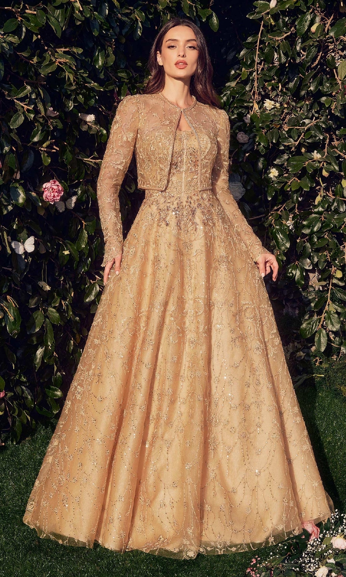 A woman stands outdoors in the Andrea & Leo MA104, a long sleeve formal dress with a matching jacket, surrounded by greenery and flowers.